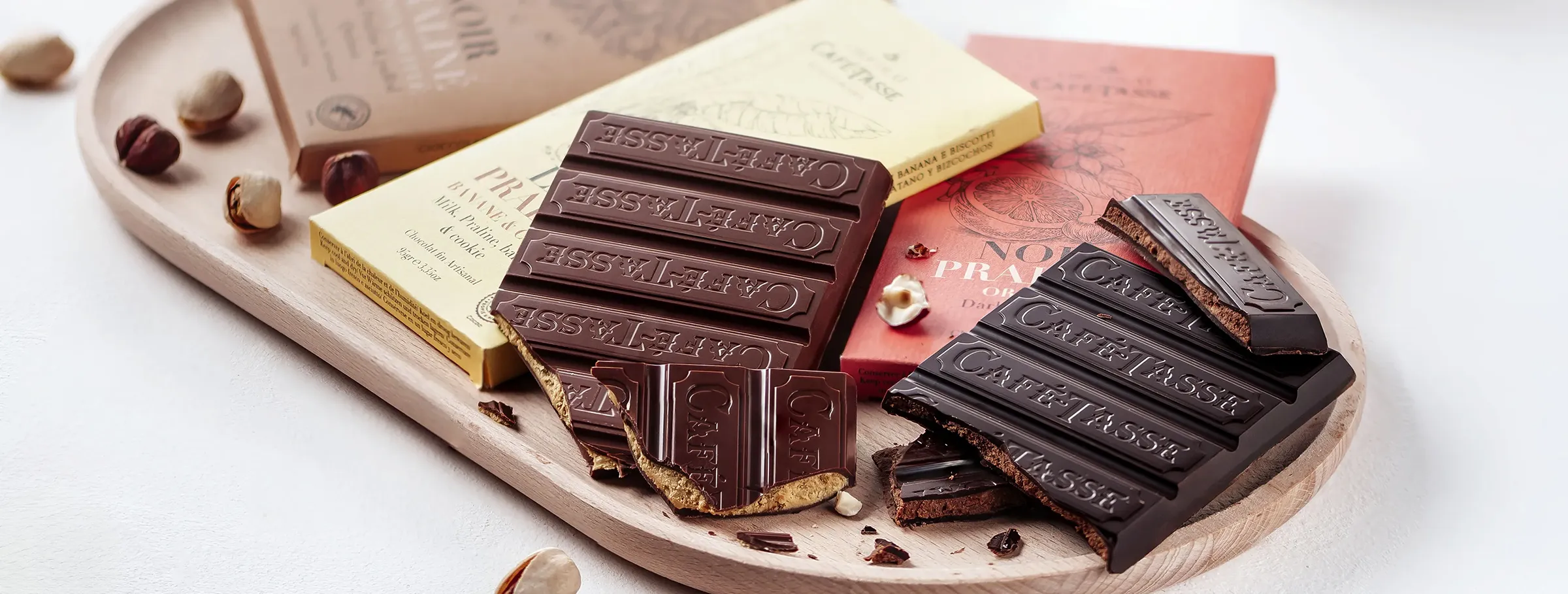 Plusieurs tablettes de chocolat Café-Tassé brisées disposées sur un plateau en bois, entourées de noisettes, aux emballages jaune, rose et beige.