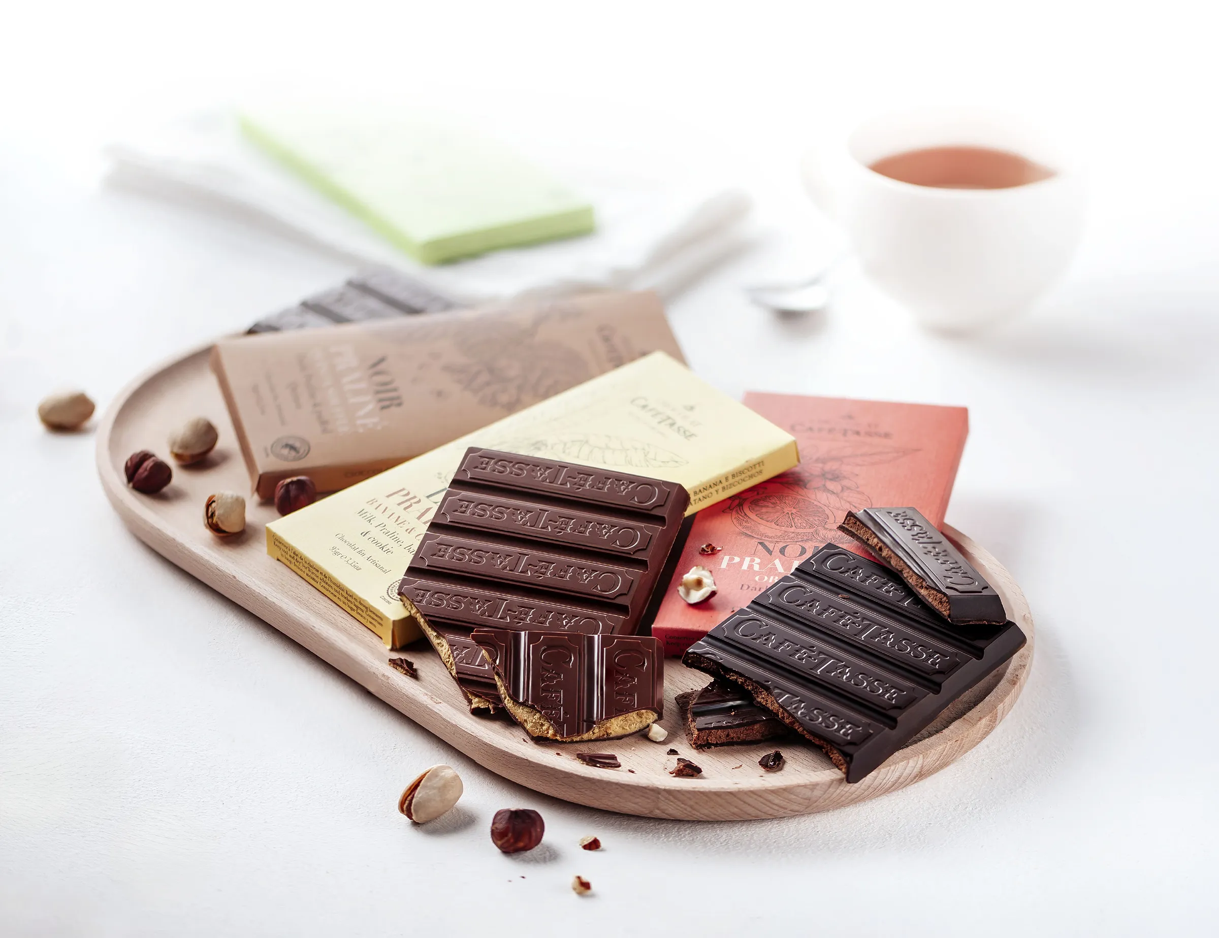 Plateau en bois avec plusieurs tablettes de chocolat Café-Tassé brisées, entourées de noisettes, une tasse de café floutée en arrière-plan.