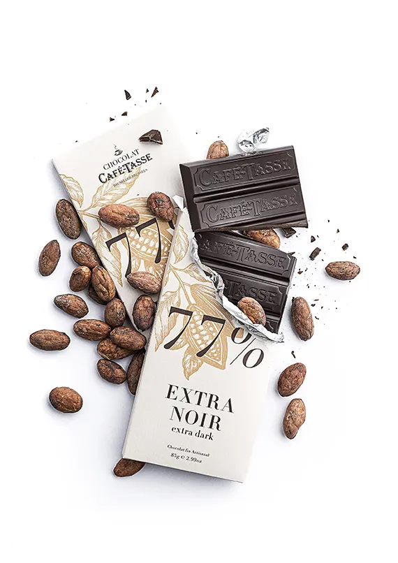 Tablette Café-Tassé « Chocolat Extra Noir » 77% brisée, emballage crème déchiré, carrés de chocolat noir entourés de fèves de cacao brutes éparpillées.