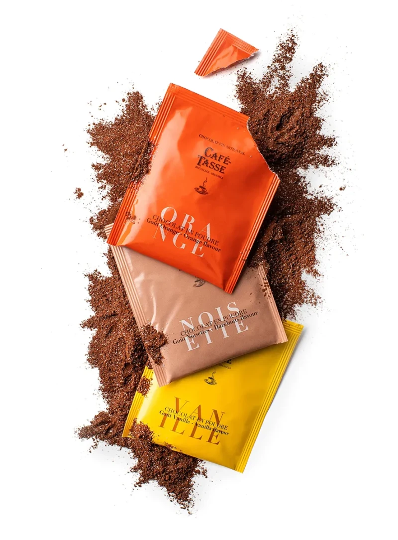 Trois sachets « Café-Tassé » de chocolat en poudre disposés en éventail sur du cacao en poudre renversé, saveurs Orange, Noisette et Vanille, emballages orange, beige et jaune.