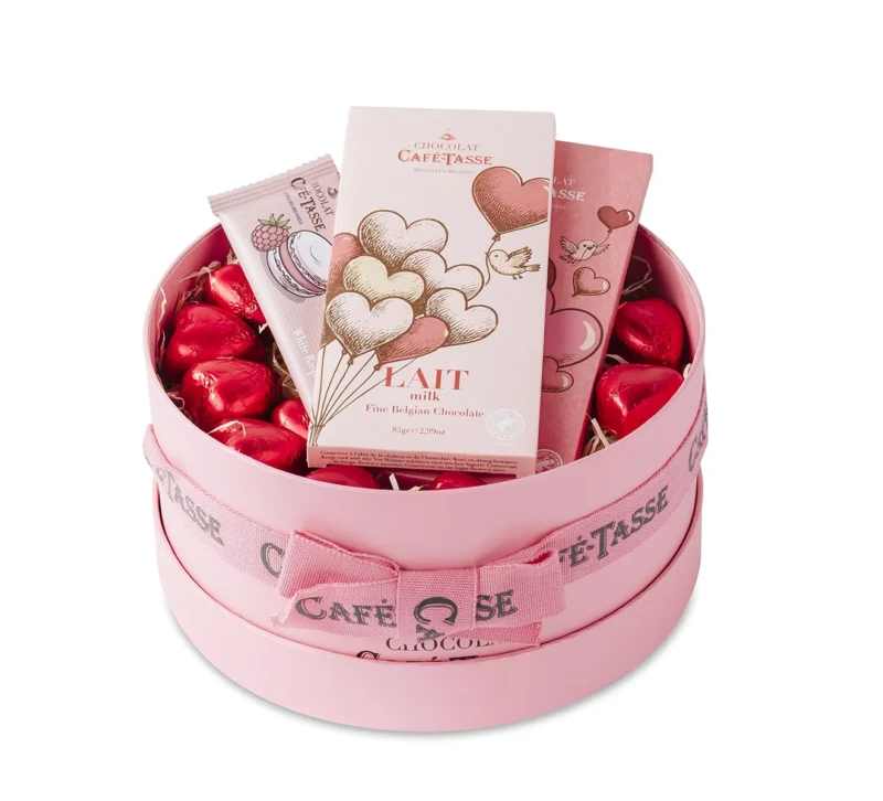 Coffret rond rose Saint-Valentin garni de cœurs en chocolat sous papillote rouge, tablette « Café-Tassé Lait » 85 gr illustrée de ballons cœurs, accompagnée d'un tube et d'une barre chocolatée roses.