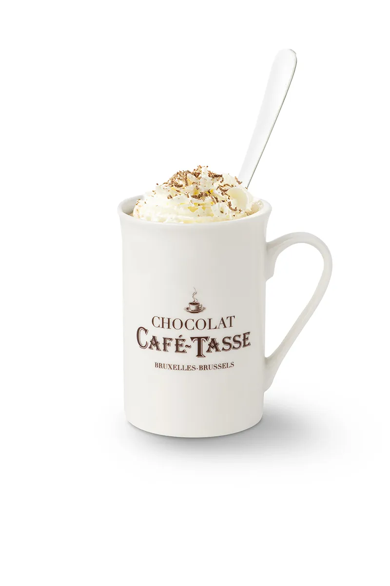 Mug blanc « Chocolat Café-Tassé Bruxelles-Brussels » rempli de chocolat chaud, garni de crème fouettée et de copeaux de chocolat râpé, cuillère plantée dans la mousse.