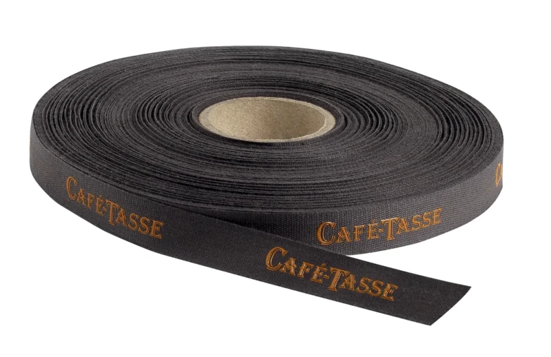 Rouleau de ruban noir posé à plat, extrémité déroulée au premier plan, tissé du mot « Café-Tassé » en lettres dorées répété en diagonale, ruban 15mm 50m.