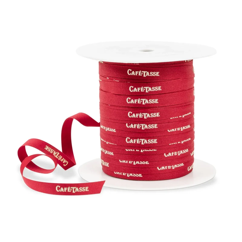 Bobine de ruban textile rouge tissé du mot « Café-Tassé » en lettres blanches répété sur toute la longueur, sur mandrin blanc, extrémité déroulée en avant.