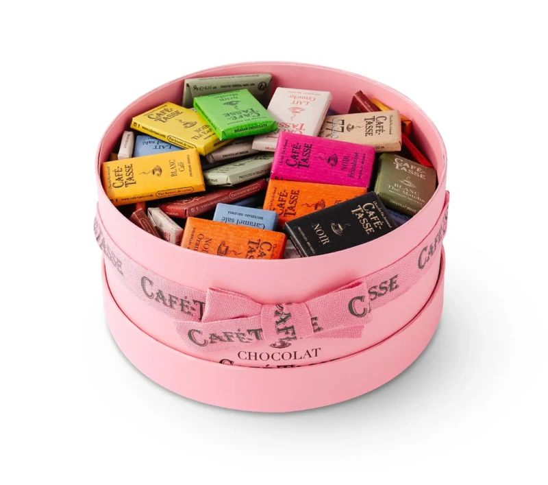 Corbeille ronde rose Café-Tassé, texte gravé sur le pourtour, remplie de 100 mini tablettes multicolores aux emballages variés (vert, jaune, rose, blanc, noir).