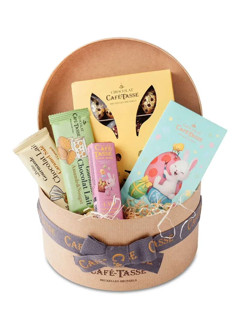 Corbeille ronde kraft nouée d'un ruban « Café-Tassé », garnie de frisure de papier, assortiment de Pâques : oreilles en chocolat, boîte illustrée d'un lapin peintre, bâtonnets et sachets aux saveurs variées.